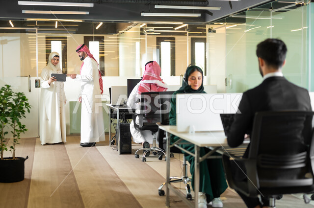 تعابير الاندماج والسرور،  بيئة العمل المريحة في الشركات السعودية، المشاركة والتعاون في إنجاز الأعمال، رجل عربي خليجي سعودي يرتدي الثوب والشماغ التقليدي مع إمرأة محجبة ترتدي العباءة يقفون داخل مقر الشركة، مهن ووظائف إدارية مكتبية