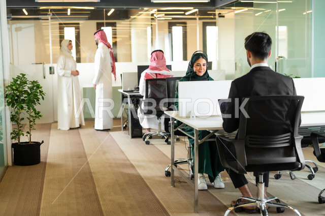 التعاون في إنجاز الأعمال، ايماءات الاندماج والسرور،  بيئة العمل المريحة في الشركات السعودية، رجل عربي خليجي سعودي يرتدي الثوب والشماغ التقليدي مع إمرأة محجبة ترتدي العباءة يقفون داخل مقر الشركة، مهن ووظائف إدارية مكتبية