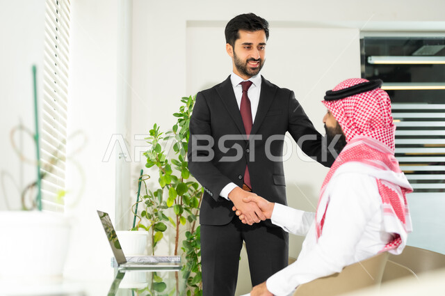 التعاون في إنجاز الأعمال، رجل عربي خليجي سعودي يرتدي الثوب والشماغ التقليدي مع رجل بالبدلة الرسمية يتصافحان بإيماءات السرور بنجاح الصفقة، الوحدة والتفاعل الجماعي لتحقيق أهداف الشركة وحل المشكلات، مهن و وظائف مكتبية، الترحيب والسلام وإلقاء التحية