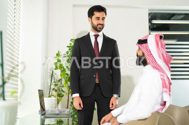 إنجاز المهام عبر الحاسوب المحمول، مهن ووظائف مكتبية سعودية، تحقيق أهداف الشركة، مفهوم إدارة الأعمال، مناقشة المشاريع ومتابعة إستراتيجية العمل، رجل عربي خليجي سعودي يرتدي الثوب والشماغ التقليدي مع رجل بالبدلة الرسمية في غرفة الاجتماعات