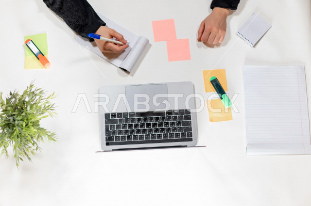 تصوير مقرب من الأعلى , لمساعدة مدير خليجية تكتب المواعيد على ورق ملاحظات أمام اللابتوب على مكتب أبيض , وظيفه ومهن , بيئة عمل , خلفية بيضاء .