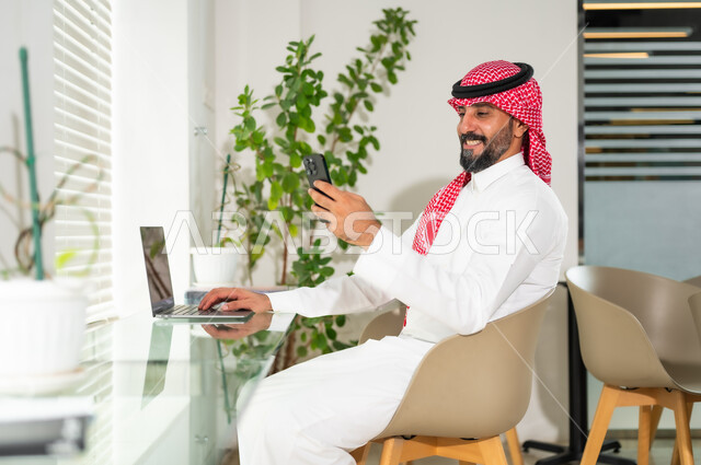مهن ووظائف ادارية مكتبية، دمج العمل مع التقنية والتكنولوجيا، إدارة شؤون الأعمال وتنظيمها عبر الاجهزة التقنية، رجل اعمال عربي خليجي سعودي مبتسم يرتدي الزي التقليدي والشماغ يمسك بيده الهاتف المحمول، العمل على الحاسوب المحمول داخل المكتب