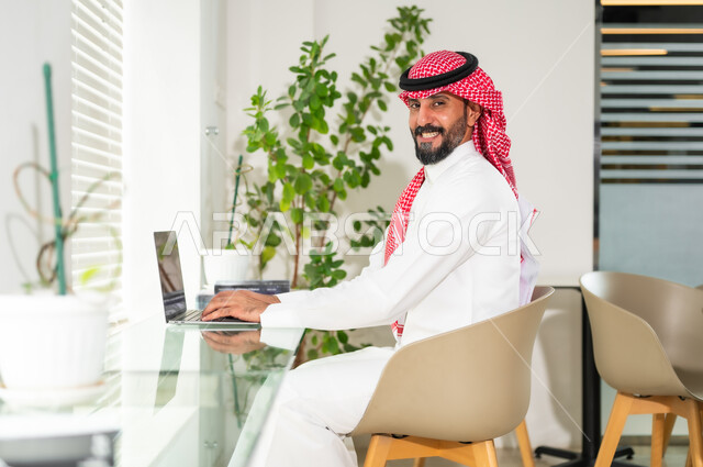 النظر إلى الكاميرا بإيماءات السرور، دمج العمل مع التقنية والتكنولوجيا، مهن ووظائف ادارية مكتبية، إدارة شؤون الأعمال وتنظيمها عبر الاجهزة التقنية، رجل اعمال عربي خليجي سعودي يرتدي الزي التقليدي والشماغ يمسك بيده الهاتف المحمول، العمل على الحاسوب المحمول