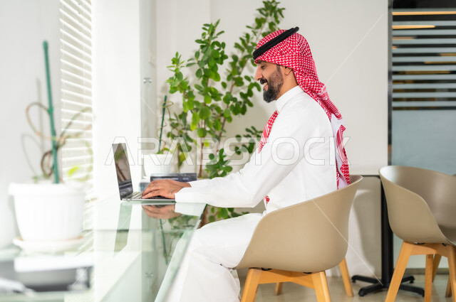 دمج العمل مع التقنية والتكنولوجيا، مهن ووظائف ادارية مكتبية، إدارة شؤون الأعمال وتنظيمها عبر الاجهزة التقنية، رجل اعمال عربي خليجي سعودي يرتدي الزي التقليدي والشماغ يمسك بيده الهاتف المحمول، العمل على الحاسوب المحمول داخل المكتب