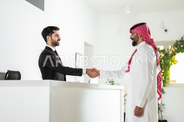 الاستعلام والاستفسار في مكتب الاستقبال في الشركات الراقية في المملكة العربية السعودية, الترحيب والسلام وإلقاء التحية بإيماءات الرضا, صورة من الجانب لرجل عربي خليجي سعودي يرتدي الثوب التقليدي والشماغ يقوم بمصافحة موظف خدمة العملاء