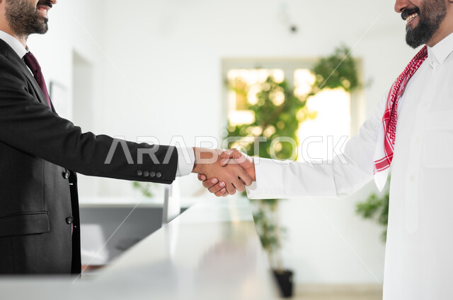 Welcome, peace and greetings, a picture from the side of a Saudi Gulf Arab man wearing the traditional thobe and shemagh shaking hands with a customer service employee, inquiries and inquiries at the reception desk in high-end companies in the Kingdom of Saudi Arabia