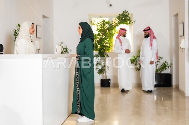 النقاش والحوار مع موظفي الشركة, صورة من الجانب لإمرأة عربية خليجية سعودية ترتدي العباءة الخضراء تقف مع موظفة الاستقبال في مكتب خدمة العملاء, مهن ووظائف نسائية, العمل في الشركات السعودية الراقية