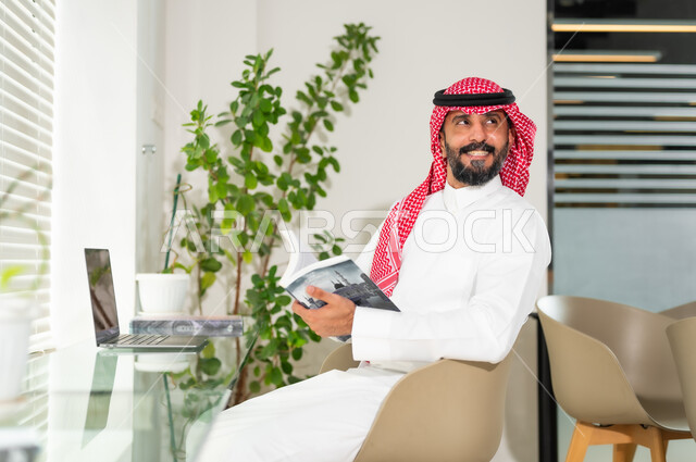 الاستمتاع بقراءة الكتب، مهن ووظائف ادارية مكتبية، إدارة شؤون الأعمال وتنظيمها عبر الاجهزة التقنية، رجل اعمال عربي خليجي سعودي مبتسم يرتدي الزي التقليدي والشماغ، العمل على الحاسوب المحمول داخل المكتب