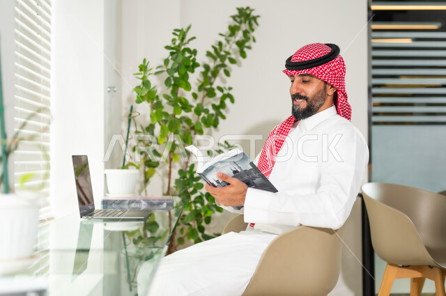 إدارة شؤون الأعمال وتنظيمها عبر الاجهزة التقنية، الاستمتاع بقراءة الكتب، مهن ووظائف ادارية مكتبية، رجل اعمال عربي خليجي سعودي يرتدي الزي التقليدي والشماغ، العمل على الحاسوب المحمول داخل المكتب