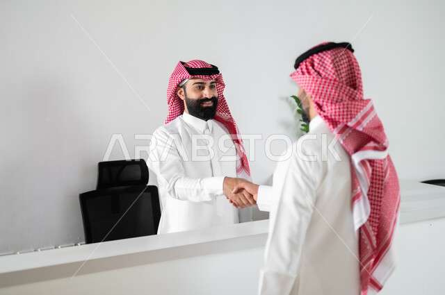 وظائف ومهن مكتبية, التحية والسلام وإبرام اتفاقيات عمل ناجحة, صورة من الخلف لرجلين عربيين خليجين سعوديين يرتديان الثوب التقليدي والشماغ يتصافحان في مقر الشركة, العمل في الشركات السعودية الراقية