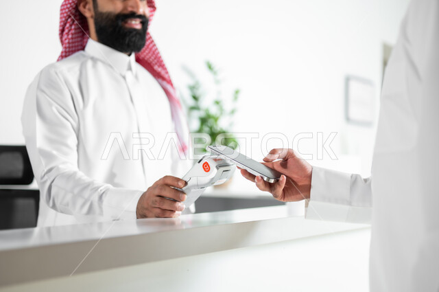 Ease of payment, reservation and inquiry procedures, customer service in tourist hotels, a close-up photo of the hands of two Saudi Gulf Arab men wearing traditional thobes and shemaghs making payment electronically using a bank credit card at the reception desk.