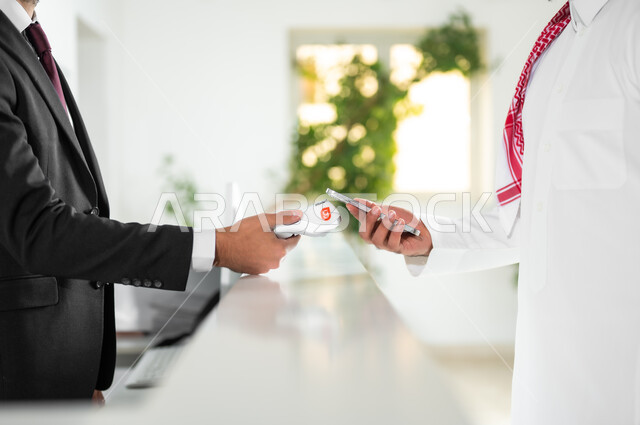إجراءات الحجز والاستفسار وسهولة الدفع, خدمة العملاء في الفنادق السياحية, صورة مقربة ليد رجل عربي خليجي سعودي يرتدي الثوب التقليدي والشماغ يقوم بالدفع بطريقة الكترونية بإستخدام الهاتف المحمول