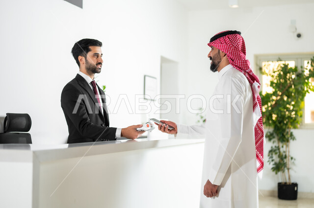 إجراءات الحجز والاستفسار وسهولة الدفع, خدمة العملاء في الفنادق السياحية, صورة من الجانب لرجل عربي خليجي سعودي يرتدي الثوب التقليدي والشماغ يقوم بالدفع بطريقة الكترونية بإستخدام الهاتف المحمول
