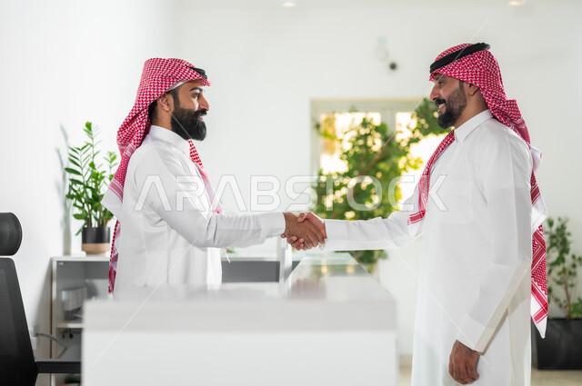 العمل في الشركات السعودية الراقية, مهن ووظائف مكتبية, التحية والسلام وإبرام اتفاقيات عمل ناجحة, صورة من الجانب لرجلين عربيين خليجين سعوديين يرتديان الثوب التقليدي والشماغ يتصافحان بتعابير السرور والرضا