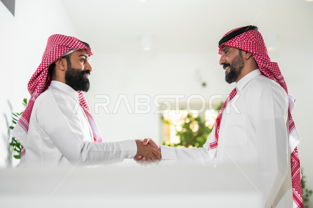 مهن ووظائف مكتبية, التحية والسلام وإبرام اتفاقيات عمل ناجحة, صورة من الجانب لرجلين عربيين خليجين سعوديين يرتديان الثوب التقليدي والشماغ يتصافحان بتعابير السرور والرضا, العمل في الشركات السعودية الراقية