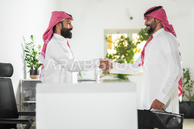 التحية والسلام وإبرام اتفاقيات عمل ناجحة, مهن ووظائف مكتبية, صورة من الجانب لرجلين عربيين خليجين سعوديين يرتديان الثوب التقليدي والشماغ يتصافحان بتعابير السرور والرضا, العمل في الشركات السعودية الراقية
