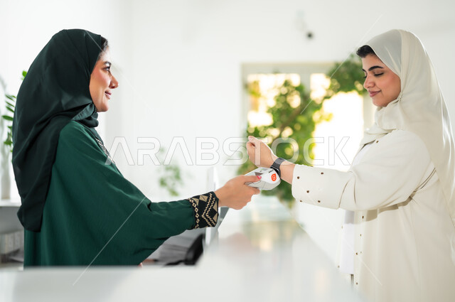 استخدام الوسائل والتقنيات الحديثة في الدفع الالكتروني, صورة مقربة من الجانب لشابة عربية خليجية سعودية ترتدي العباءة والحجاب تقف في مكتب الاستقبال, سهولة الدفع الالي من خلال ساعة السمارت عبر الهاتف المحمول, مهن ووظائف نسائية في الشركات الراقية