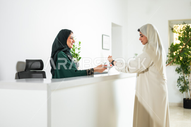 استخدام الوسائل والتقنيات الحديثة في الدفع الالكتروني, صورة من الجانب لشابة عربية خليجية سعودية ترتدي العباءة والحجاب تقف في مكتب الاستقبال, سهولة الدفع الالي من خلال ساعة السمارت عبر الهاتف المحمول, مهن ووظائف نسائية في الشركات الراقية