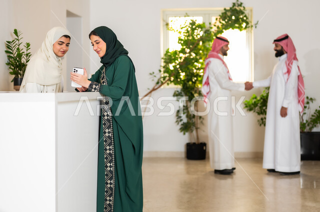 استخدام الأجهزة والتقنيات الحديثة, صورة من الجانب لإمرأة عربية خليجية سعودية ترتدي العباءة الملونة تمسك بيدها الهاتف المحمول وتقوم بالحوار مع موظفة الاستقبال في إحدى الشركات السعودية الراقية, التشاور والسؤال في مكتب خدمة العملاء