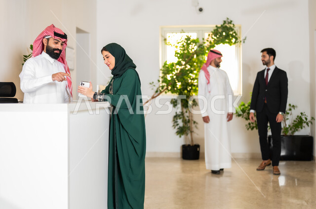 استخدام الأجهزة والتقنيات الحديثة, شابة عربية خليجية سعودية ترتدي عباءة ملونة تمسك بيدها الهاتف المحمول وتقف عند مكتب الاستقبال, المناقشة والحوار حول أهداف الشركة, بيئة العمل في الشركات الخليجية, رفع إصبع السبابة والإشارة إلى الجوال