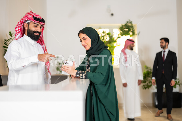 رفع إصبع السبابة والإشارة إلى الجوال, استخدام الأجهزة والتقنيات الحديثة, شابة عربية خليجية سعودية ترتدي عباءة ملونة تمسك بيدها الهاتف المحمول وتقف عند مكتب الاستقبال, المناقشة والحوار حول أهداف الشركة, بيئة العمل في الشركات الخليجية