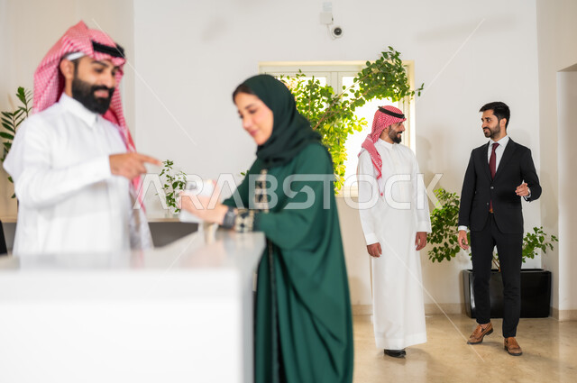 The work environment in Gulf companies, raising the index finger and pointing at the mobile phone, using modern devices and technologies, a young Saudi Gulf Arab woman wearing a colorful abaya, holding a mobile phone in her hand and standing at the reception desk, discussion and dialogue about the company’s goals