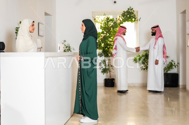 المصافحة والاتفاق بتعابير التحية والسلام, الاستفسار حول خدمة العملاء, صورة من الجانب لشابة عربية خليجية سعودية ترتدي العباءة الخضراء تقوم بالنقاش والسؤال مع موظفة الاستقبال,  العمل في الشركات السعودية الراقية