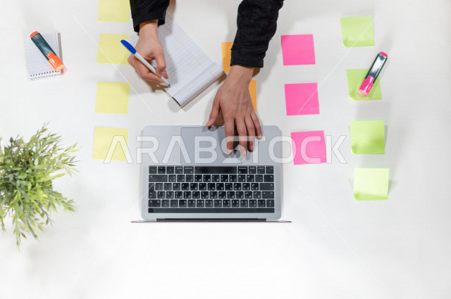  تصوير مقرب من الأعلى , لمساعدة مدير خليجية تكتب المواعيد على ورق ملاحظات أمام اللابتوب على مكتب أبيض , وظيفه ومهن , بيئة عمل , خلفية بيضاء .