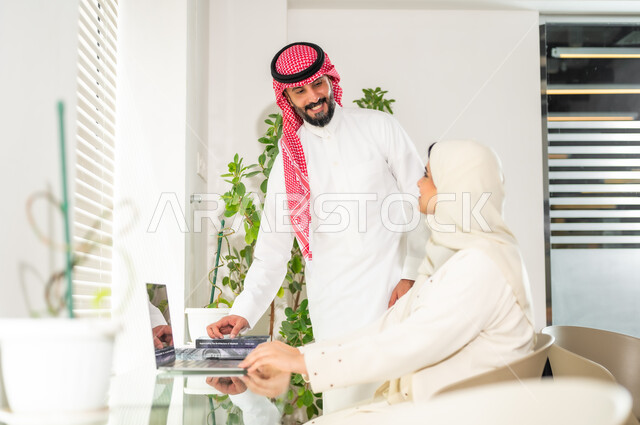 إنجاز المهام عبر الحاسوب المحمول، مهن ووظائف مكتبية سعودية، تحقيق أهداف الشركة، مفهوم إدارة الأعمال، مناقشة المشاريع ومتابعة إستراتيجية العمل، رجل عربي خليجي سعودي يرتدي الثوب والشماغ التقليدي مع إمرأة محجبة ترتدي العباءة  في غرفة الاجتماعات