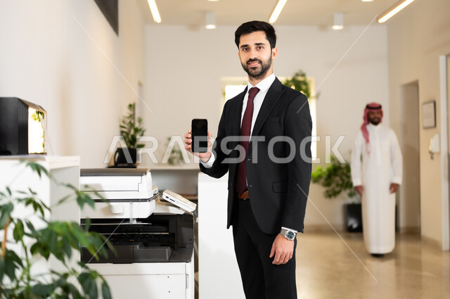 عرض شاشة سوداء فارغة عبر الهاتف المحمول, استخدام الأجهزة والتقنيات الحديثة والمتطورة, عقد اتفاقيات عمل ناجحة, رجل أعمال عربي خليجي سعودي يرتدي الزي الرسمي ينظر إلى الكاميرا بتعابير السرور, العمل في الشركات الخليجية الراقية