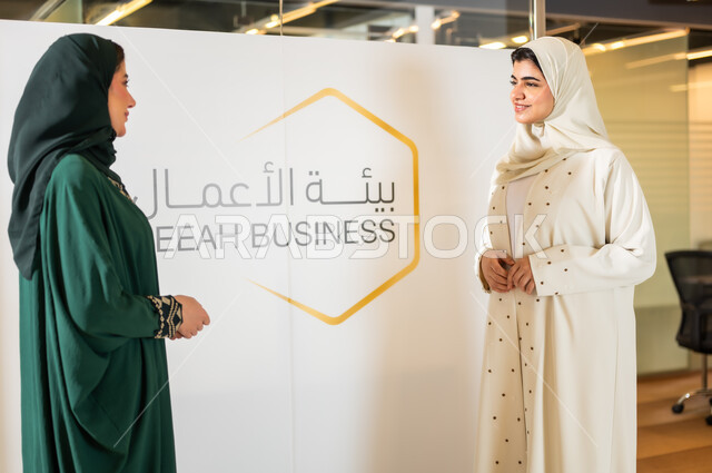 Discussions and dialogues about the concept of business management, two veiled Saudi Gulf Arab businesswomen wearing modern abayas, exchanging conversations about office work, answering questions and inquiries in the educational conference.