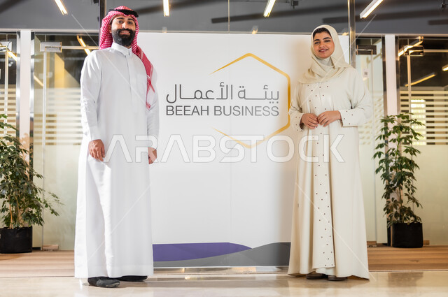 Saudi administrative professions and jobs, educational seminars on the labor environment and market, a Saudi Arabian Gulf businessman in traditional dress standing upright and looking at the camera, a veiled employee wearing a modern beige abaya, conferences to explain the various areas of office work in companies.