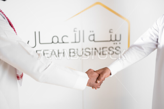 المناقشات والمحادثات عن مفهوم ادارة العمل، الرد على الأسئلة والاستفسارات في المؤتمر التثقيفي، تبادل الأحاديث حول الأعمال المكتبية، صورة مقربة ليدان رجلا أعمال عربيان خليجيان سعوديان يرتديان الزي التقليدي يقومان بمصافحة بعضهما