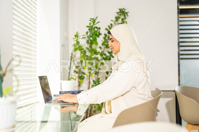 استخدام الوسائل التقنية الحديثة والمتطورة لإنجاز المهام، مهن ووظائف مكتبية نسائية, إدارة وتنظيم الأعمال ضمن مقر الشركة, امرأة عربية خليجية سعودية محجبة ترتدي العباءة تعمل على الحاسب المحمول, بيئة العمل المريحة في الشركات السعودية