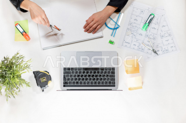  تصوير مقرب من الأعلى , لمهندسة تصميم داخلي عربية سعودية , مهندسة تصمم الأثاث الداخلي أمام اللابتوب على مكتب أبيض , وظيفه ومهن , بيئة عمل , خلفية بيضاء .