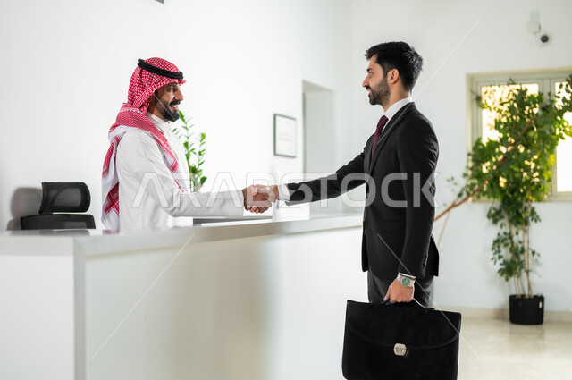 Office jobs and professions, agreeing on projects and business plans, shaking hands, greetings and greetings with expressions of pleasure, a picture from the side of a Saudi Arabian Gulf businessman wearing a formal uniform shaking hands with an employee in the company.