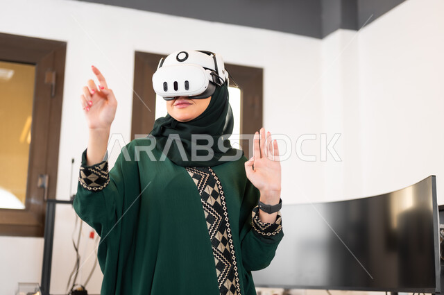 ايماءات وتعابير الاندماج والتركيز في الواقع الافتراضي، التقدم التقني و التحول الرقمي، استخدام الأجهزة والتفنيات الحديثة المتطورة، محاكاة العالم الخيالي، سيدة عربية خليجية سعودية محجبة ترتدي نظارات ثلاثية الأبعاد تقف بداخل مكتبها وتقوم بالإشارة الى شيء ما