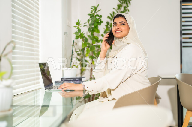 Business management and communication with customers via mobile phone, a close-up image of a smiling Saudi Gulf Arab woman wearing a gray abaya using the mobile phone with cheerful and happy gestures, completing office tasks at the company headquarters, women’s professions and jobs, the Saudi work environment