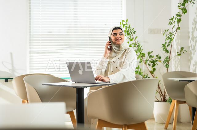 The Saudi work environment, completing office tasks at the company’s headquarters, managing business and communicating with customers via mobile phone, a smiling Saudi Gulf Arab woman wearing an abaya using the mobile phone with gestures of joy and happiness, women’s professions and jobs