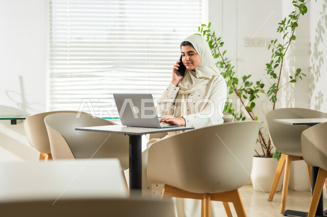 دمج التقنية والتكنولوجيا في الحياة العملية، إنجاز المهام المكتبية في مقر الشركة، إدارة الأعمال والتواصل مع العملاء عبر الجوال, سيدة عربية خليجية سعودية مبتسمة ترتدي العباءة تقوم باستخدام الهاتف المحمول بإيماءات البشاشة والسرور, مهن ووظائف نسائية