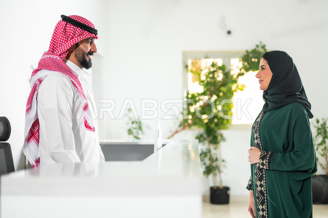 بيئة العمل المريحة في الشركات الراقية. البشاشة وحسن الاستقبال, الحوار والنقاش وتقديم الخدمات للعملاء, صورة من الجانب لشابة عربية خليجية سعودية ترتدي العباءة الخضراء والحجاب تقوم بالاستفسار في مكتب الخدمة, مهن ووظائف سعودية