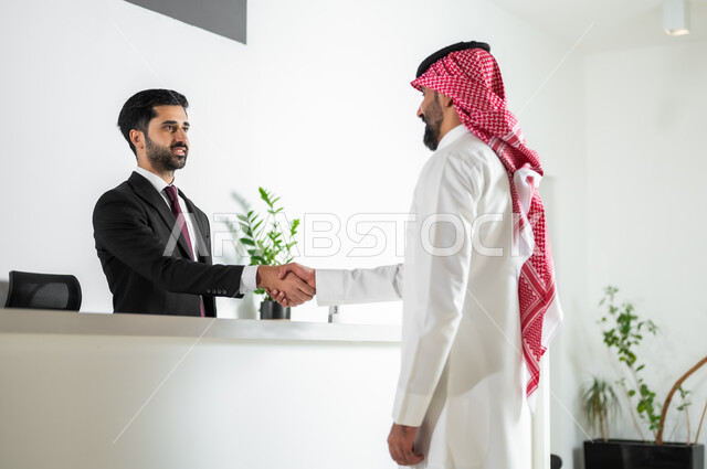 السلام والترحيب وإلقاء التحية, صورة من الخلف لرجل عربي خليجي سعودي يرتدي الثوب التقليدي والشماغ يقوم بمصافحة موظف خدمة العملاء, الاستعلام والاستفسار في مكتب الاستقبال في الشركات الراقية في المملكة العربية السعودية