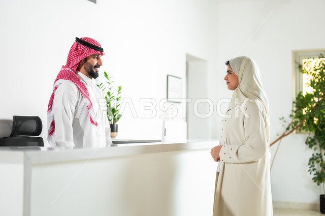 بيئة العمل المريحة في الشركات الراقية, البشاشة وحسن الاستقبال, الحوار والنقاش وتقديم الخدمات للعملاء, صورة من الجانب لشابة عربية خليجية سعودية ترتدي العباءة والحجاب تقوم بالاستفسار في مكتب الخدمة, مهن ووظائف سعودية