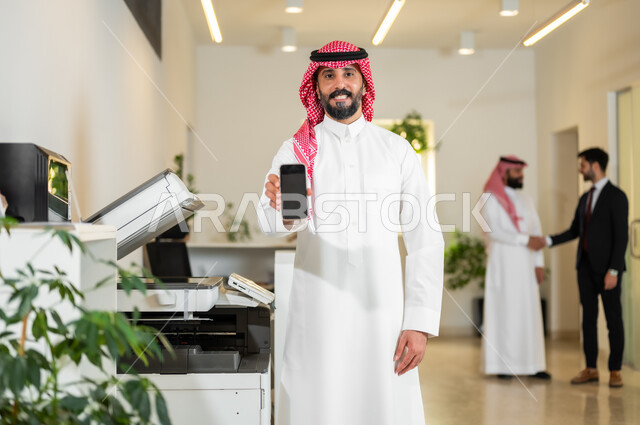 استخدام الأجهزة والتقنيات الحديثة والمتطورة, عرض شاشة سوداء فارغة عبر الهاتف المحمول, عقد اتفاقيات عمل ناجحة, رجل أعمال عربي خليجي سعودي يرتدي الثوب التقليدي والشماغ ينظر إلى الكاميرا بتعابير السرور, العمل في الشركات الخليجية الراقية