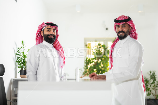 التعاون ومفهوم إدارة الأعمال المكتبية, موظفان عربيان خليجيان سعوديان يرتديان الثوب التقليدي والشماغ يقومان بالنظر إلى الكاميرا بابتسامة, بيئة العمل الراقية في الشركات الخليجية