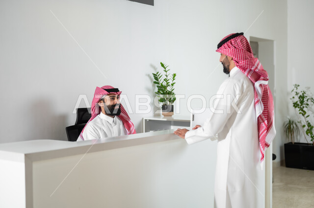 مهن ووظائف إدارية, العمل في الشركات الخليجية الراقية, الحوار والنقاش مع موظف خدمة الزبائن, صورة من الخلف لرجل عربي خليجي سعودي يرتدي الثوب التقليدي والشماغ يقوم بالاستفسار والسؤال في مكتب الخدمات, البشاشة وحسن استقبال الزوار