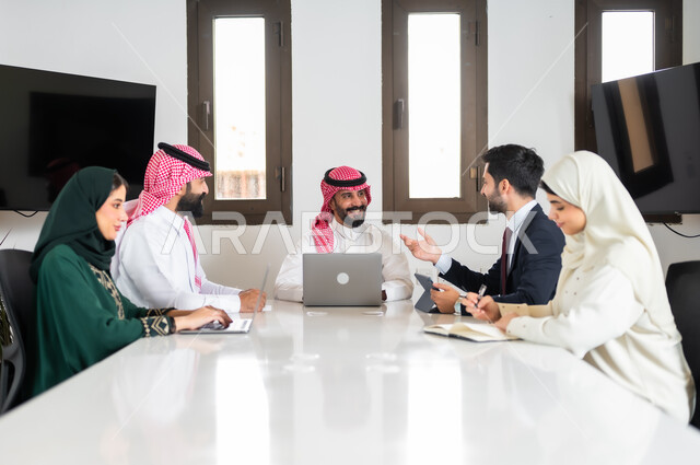 بيئة العمل المريحة في الشركات السعودية, اجتماع عمل بين موظفين عرب خليجيين سعوديين في غرفة الاجتماعات، ايماءات السعادة والسرور لبدء الشراكة، نقاشات خاصة بالعمل,التفاهم والتعاون بين الزملاء لتحقيق أهداف الشركة, استخدام الأجهزة التقنية الحديثة في المكاتب