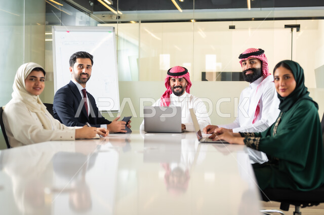 النظر الى الكاميرا بايماءات السعادة والسرور لبدء الشراكة، اجتماع عمل بين موظفين عرب خليجيين سعوديين في غرفة الاجتماعات، التفاهم والتعاون بين الزملاء لتحقيق أهداف الشركة, بيئة العمل المريحة في الشركات السعودية, استخدام الأجهزة التقنية الحديثة في المكاتب