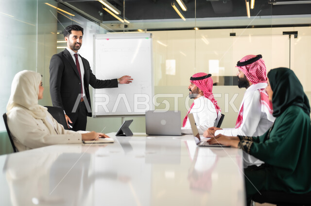 استعمال السبورة لتوضيح الأفكار، اجتماع عمل بين موظفين عرب خليجيين سعوديين في غرفة الاجتماعات، التفاهم والتعاون بين الزملاء لتحقيق أهداف الشركة, بيئة العمل المريحة في الشركات السعودية, استخدام الأجهزة التقنية الحديثة في المكاتب