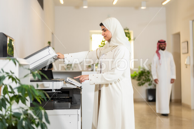 Comfortable work atmosphere in companies, female office professions and jobs, a veiled Saudi Arabian Gulf employee wearing a modern beige abaya who prints important work papers via the paper printer in the office, using modern technical devices and machines in the offices